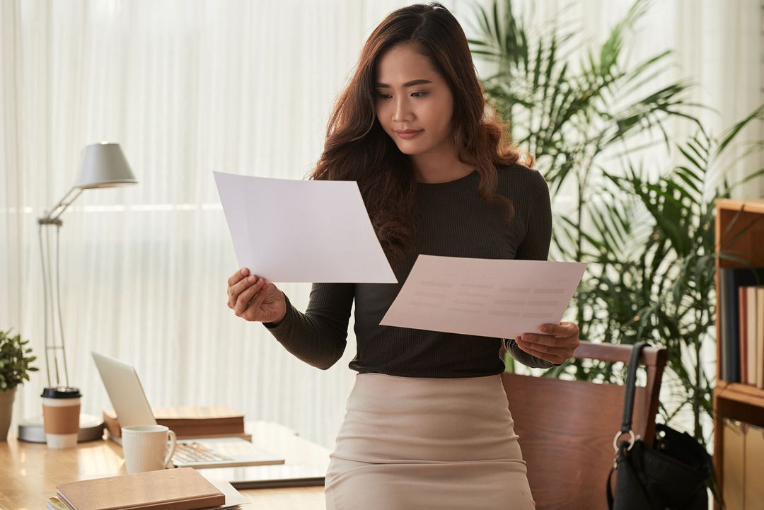 A Woman Standing And Comparing Two Pieces Of Paper